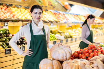 Woman seller in an apron stands in the vegetable section of a supermarket and offers a seasonal pumpkin. Grocery store worker checks pumpkin quality and invites to shop