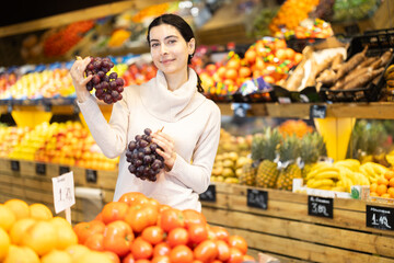Near shelves and display cases in sales area of store, woman customer takes ripe grape out of box. Wide range of agricultural products. Self-service, comfortable shopping.