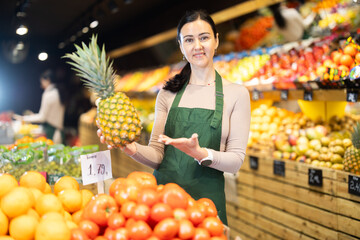 Polite middle-aged female seller offering pineapple standing by counter in large grocery store