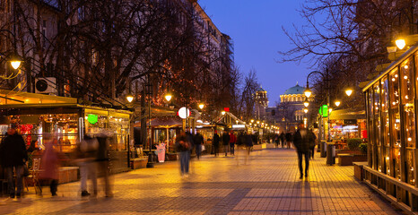 Vitosha Boulevard in Sofia sparkling at night with festive string lights decorating cafes and restaurants, creating inviting winter ambiance, and people walking towards illuminated St. Nedelya Church.