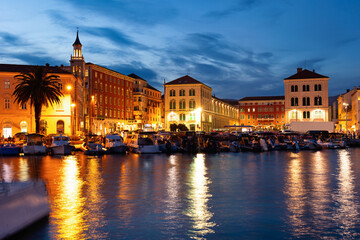 Naklejka premium Twilight and lantern light, largest port is located in Split. Private boats and yachts sway on waves, pedestrian tourists walk along embankment.
