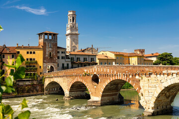 Scenic summer view of Ponte Pietra, oldest Roman bridge in Verona with graceful stone arches spanning Adige River, framed by houses draped in greenery and cathedral bell tower towering above skyline.