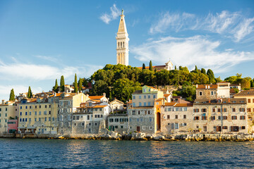 Fototapeta premium Bell tower of Church of St. Euphemia rises above city of Rovinj, citys dominant feature. Citys architecture and nature attract tourists. View of city from boat.