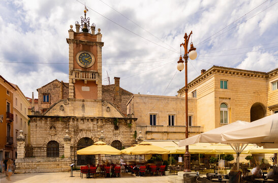 Summer scene of Narodni Trg in Zadar, Croatia, with historic City Sentinel Clock Tower rising into cloudy sky, old stone buildings, open-air cafes, and people walking across square
