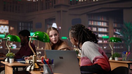 Young scholar focuses on exam preparations in the campus library with colleague, takes notes on laptop from reference books in a peaceful study setting. Girl uses the database materials. Camera A.