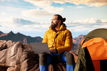 Fototapeta premium Workaholic explorer always connected and addicted to uninterrupted work, opening his laptop on the mountain top. Young hiker traveler with internet dependency enjoys strong signal.