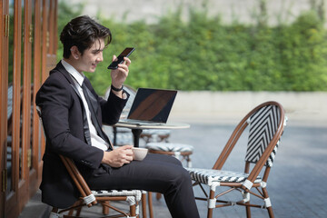 A young businessman in a black suit works at a coffee shop, using a laptop and a mobile phone while drinking coffee outdoors. The New technology business works.