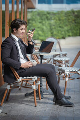 A young businessman in a black suit works at a coffee shop, using a laptop and a mobile phone while drinking coffee outdoors. The New technology business works.