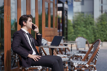 A young businessman in a black suit works at a coffee shop, using a laptop and a mobile phone while drinking coffee outdoors. The New technology business works.