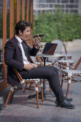 A young businessman in a black suit works at a coffee shop, using a laptop and a mobile phone while drinking coffee outdoors. The New technology business works.