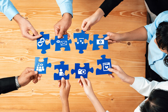 A diverse group of individuals collaborates around a wooden table, connecting blue puzzle pieces featuring social media icons, symbolizing teamwork and communication. Amity