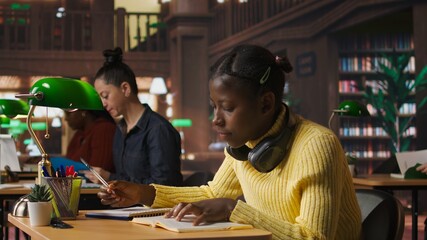 Young focused scholar doing her homework at a library desk after classes, engrossed in literature...