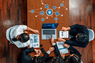 A diverse team gathers around a table for a business meeting, discussing security strategies while reviewing data charts and digital protection icons on a laptop. Amity