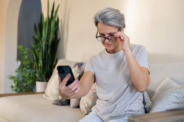 Senior woman in glasses using smartphone at home, squinting and adjusting vision