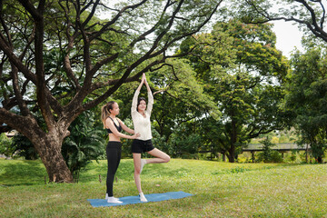 Fototapeta premium Asian woman in sportswear stretching workout outdoor on yoga mat in park with her trainer. Sporty buddy women work out together in green garden. Sporty girl engage in exercise with partner in nature.