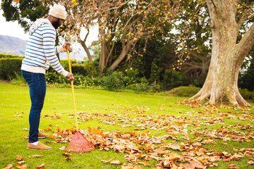 Mid-adult African American man raking leaves on lawn with yellow-handled red leaf rake, copy space