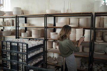Female artisan in apron checking unglazed pottery at studio shelves with tablet, copy space