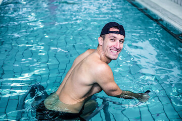 Shirtless swimmer wearing backward black cap and trunks resting on gym pool floor reflecting lights