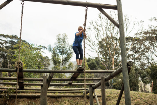 Athletic woman balancing on wooden plank at outdoor obstacle course while gripping thick rope