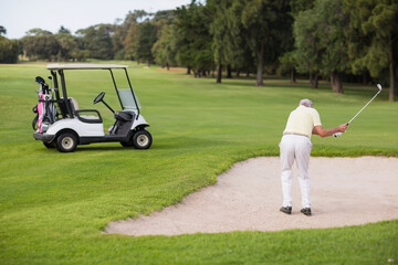 Senior man swinging golf club in sand bunker beside golf cart on golf course, copy space