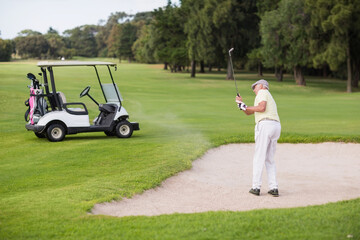 Senior man swinging club, hitting golf ball from bunker on golf course beside cart, copy space