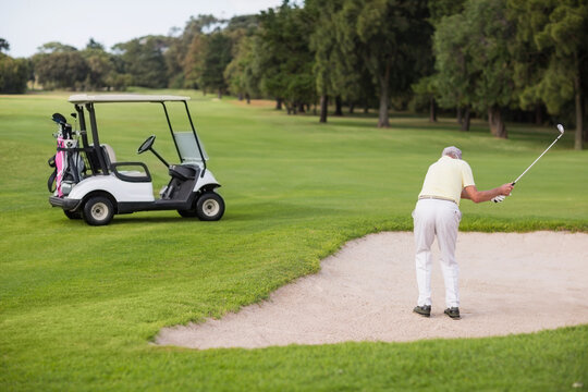 Senior man swinging golf club in sand bunker beside golf cart on golf course, copy space