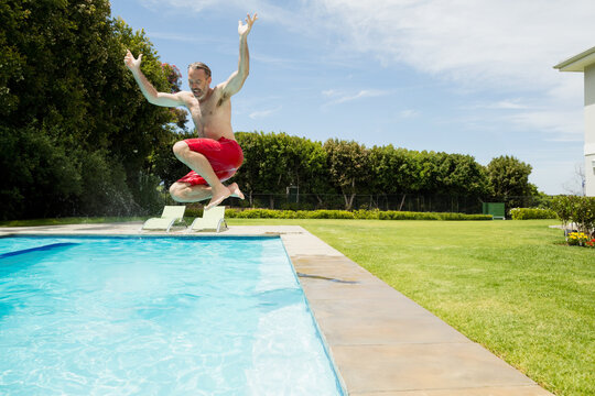 Adult male jumping into clear blue pool at backyard deck with white lounge chairs, copy space