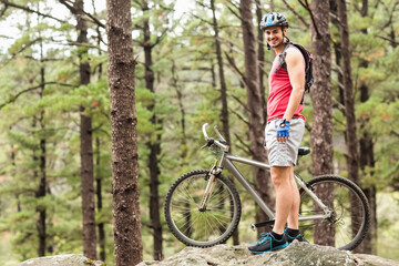 Fototapeta premium Man standing on outcrop in pine forest holding mountain bike wearing helmet backpack, copy space