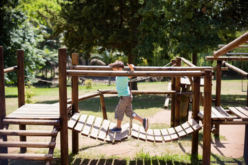 Suspended wooden plank bridge swinging over grassy ground near dirt pathways in sunny park