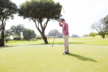 Asian man holding flagstick with white flag upright on putting green at golf course, copy space