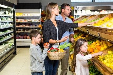 Family of four shopping in supermarket produce aisle holding wicker basket and selecting fruits