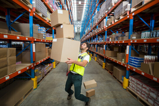 Worker wearing reflective vest balancing three boxes tipping in warehouse among blue-orange racks