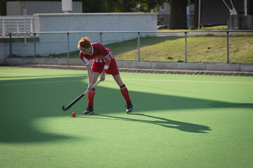 Male field hockey player bending forward, gripping stick, focusing on red ball on turf