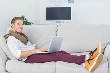 Man reclining on beige couch in living room typing on laptop with potted plant, wall notes