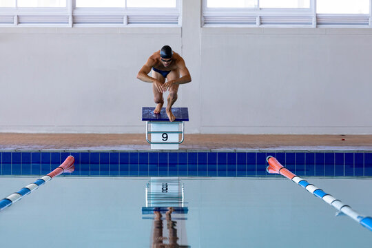 Male swimmer crouching on starting block labeled 9 at pool, wearing black swim cap and goggles