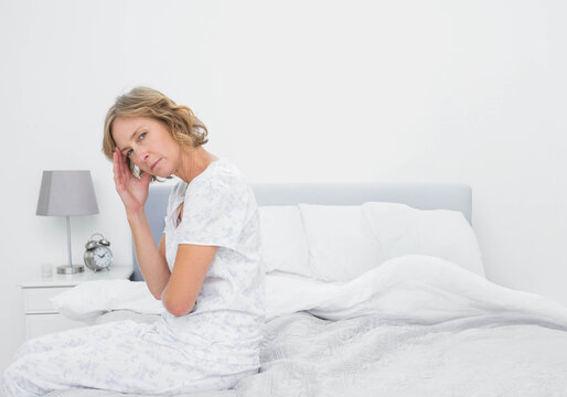 Senior woman sitting on bed in bedroom holding head with hand near nightstand lamp and clock