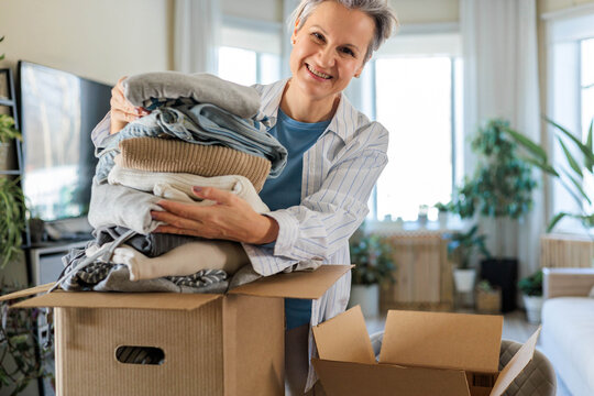 Portrait of smiling gray haired mature woman pack clothes pile to cardboard box for donation