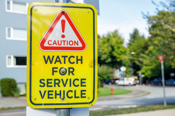 Bright yellow caution sign with red exclamation mark warning "Watch for Service Vehicle" in suburban street setting