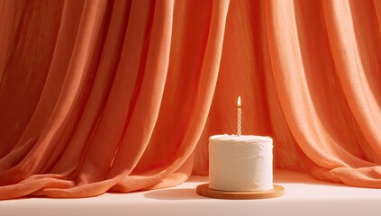A single white cake with a lit candle sits before a backdrop of soft peach-toned fabric