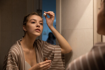 Beautiful young woman taking care of her face at home, skincare procedures, selective focus. Portrait of a woman applying a serum and doing face massage for her face in her bathroom. Real photo.