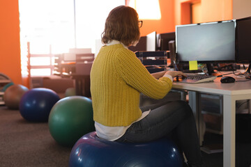 Woman balancing on stability ball and typing on keyboard at office desk under sunlight