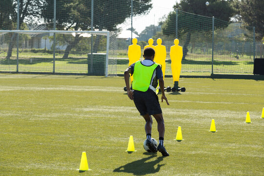 African American male soccer player dribbling soccer ball through cones past goal dummies by fence