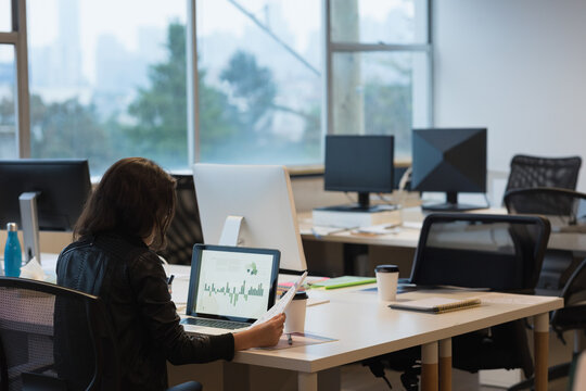 Woman reviewing charts on tablet while holding printed report at open office desk with coffee cup