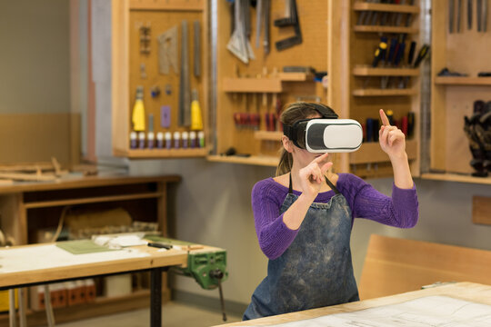 female teenager wearing VR headset, denim apron examining blueprints in woodworking workshop
