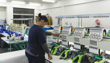 Woman adjusting multi-head embroidery machine controls inside workshop with thread spools and caps