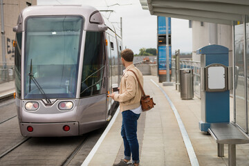 Mid-adult male commuter waiting for silver tram on platform holding coffee cup and leather satchel