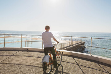 Man standing behind orange bicycle by metal railing on seaside promenade, overlooking ocean