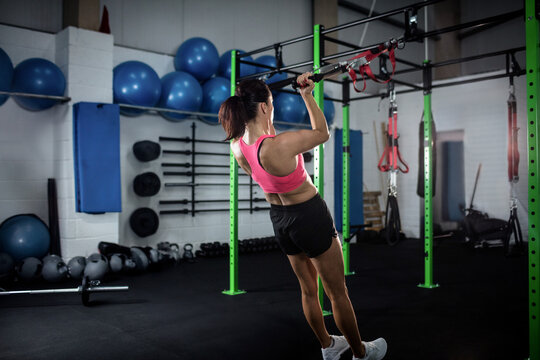 Female exerciser pulling suspension straps on green metal rig in gym with blue stability balls