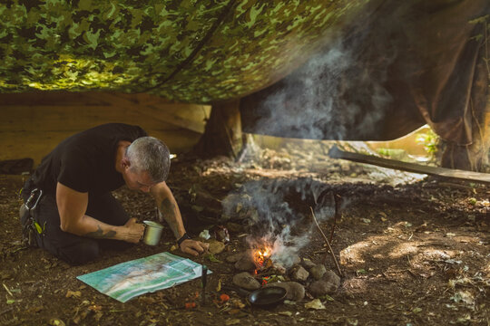 Man kneeling under camouflage tarp in woods consulting map near smoking campfire holding metal mug - Powered by Adobe