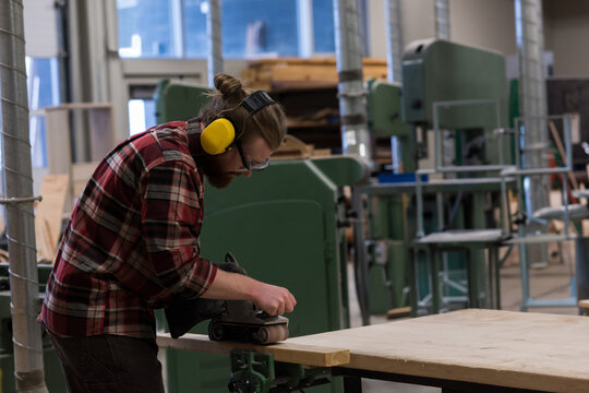 Male carpenter with goggles and earmuffs smoothing board with belt sander in workshop, copy space